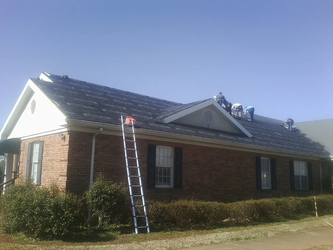 Crew tearing shingles off a brick ranch roof with a ladder set — Emergency Roof Repair work by our crew in Rindge