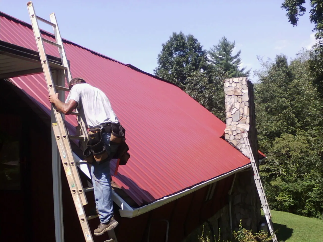 Roofer on a ladder beside a red metal roof and stone chimney — Soffit & Fascia Repair by Roofing Brothers in Rindge
